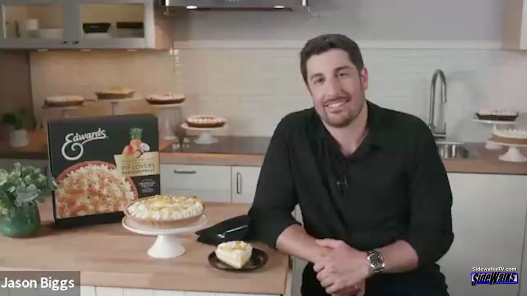 Jason Biggs smiles on a kitchen set with pies on table next to him
